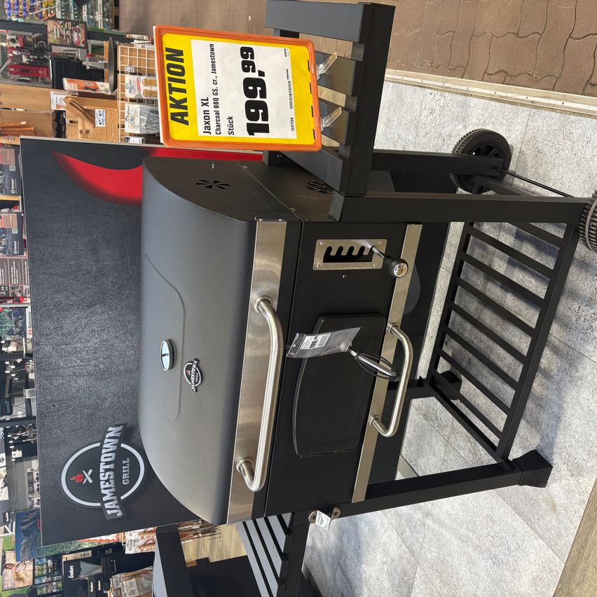 A large black and silver barbecue grill with a price tag is on display in a store. The grill has a wheel, handles, and a stainless steel lid.