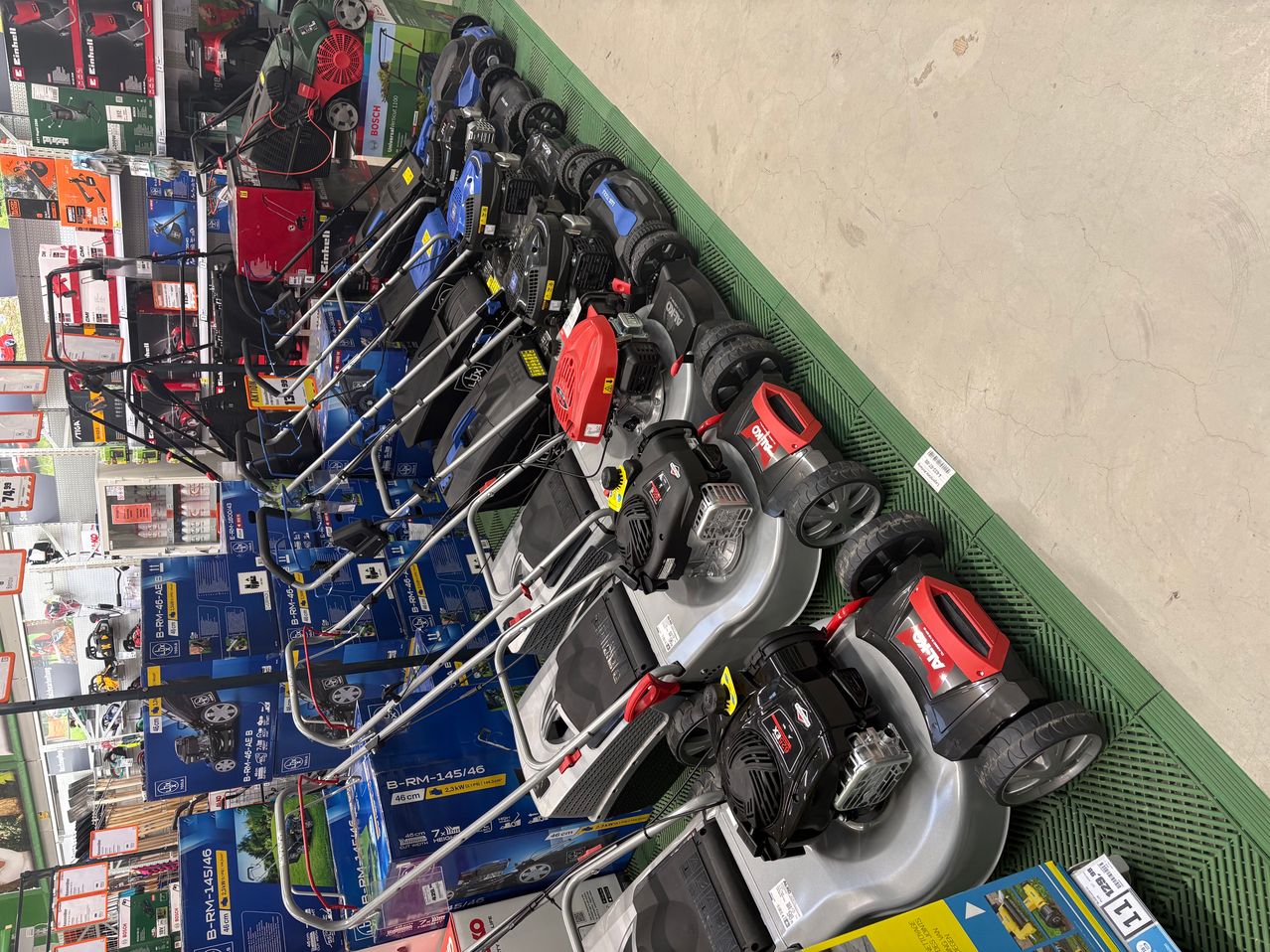 A collection of lawn mowers, primarily red and black, are neatly stacked in a store. They are placed on a green tray, and the floor is made of concrete.