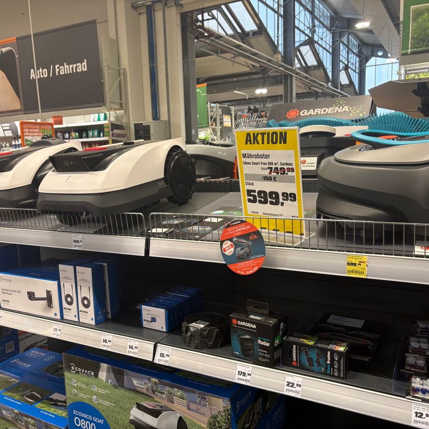A store display features robotic lawn mowers on shelves, prominently showing a price tag for 599.99 Euros. Various brands and products are visible, with an advertisement banner for Auto/Fahrrad in the background.