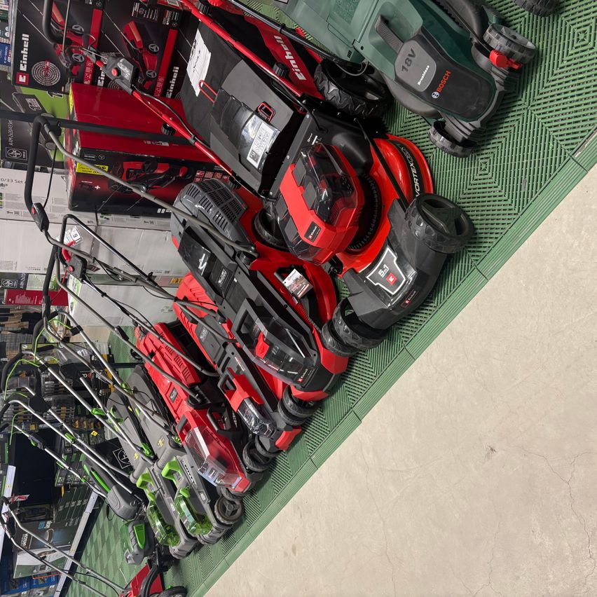A store display of various lawn mowers, mostly red and black, arranged on a green mat. Some mowers have white labels with text.
