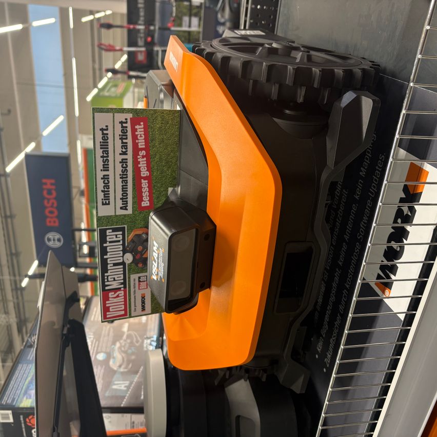 A lawnmower, orange and black, is displayed in a store. It has a green sign that reads 'Attachment not included.' The product is near a wire shelf.