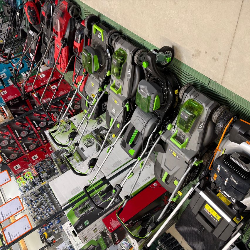 Eine Draufsicht auf verschiedene Rasenmäher in verschiedenen Farben, die an einer Ladenwand hängen. Einige sind in Kartons, andere nicht.