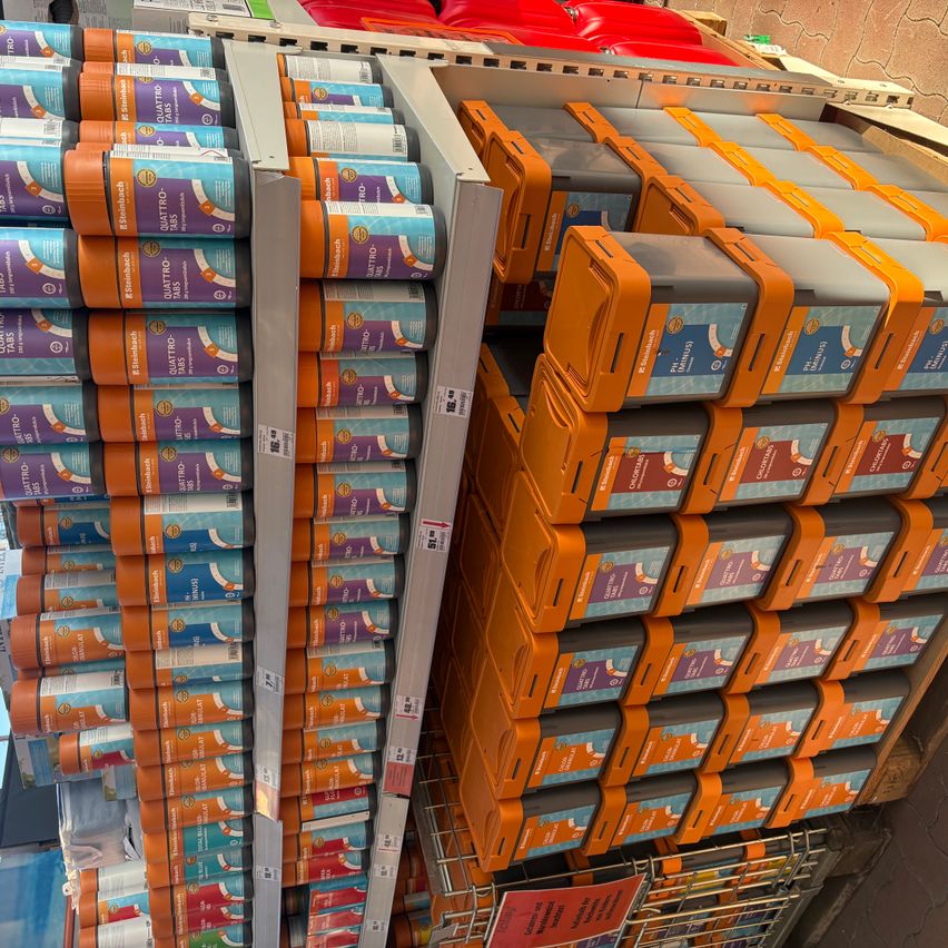 Ein Geschäft mit Regalen, die Stapel von orangefarbenen und grauen Kisten mit der Aufschrift 'Quattro' und 'Peel & Stick' anzeigen. Die Regale sind in einem Rastermuster angeordnet.