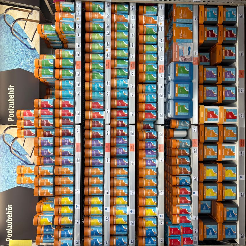 Reihen von orangefarbenen, blauen und grünen Behältern, wahrscheinlich Poolreinigungsmittel, sind auf Regalen gestapelt. Unten links befindet sich ein gelbes Schild mit dem Wort Poolzubehör, und der Hintergrund ist ein Bild eines Schwimmbeckens.
