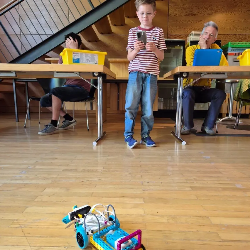 Ein junger Junge steht in einem Raum mit einem Roboter vor ihm. Zwei Erwachsene sitzen an Tischen in der Nähe, einer mit einem Laptop und der andere benutzt ein Handy.