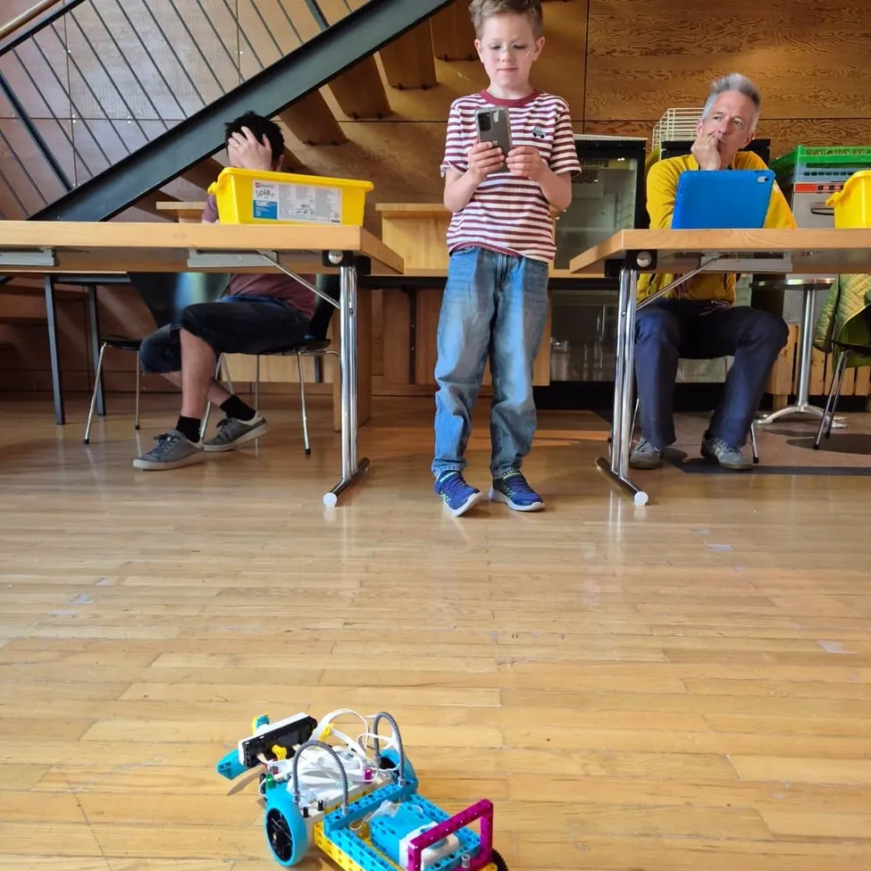 Ein junger Junge steht in einem Raum mit einem Roboter vor ihm. Zwei Erwachsene sitzen an Tischen in der Nähe, einer mit einem Laptop und der andere benutzt ein Handy.