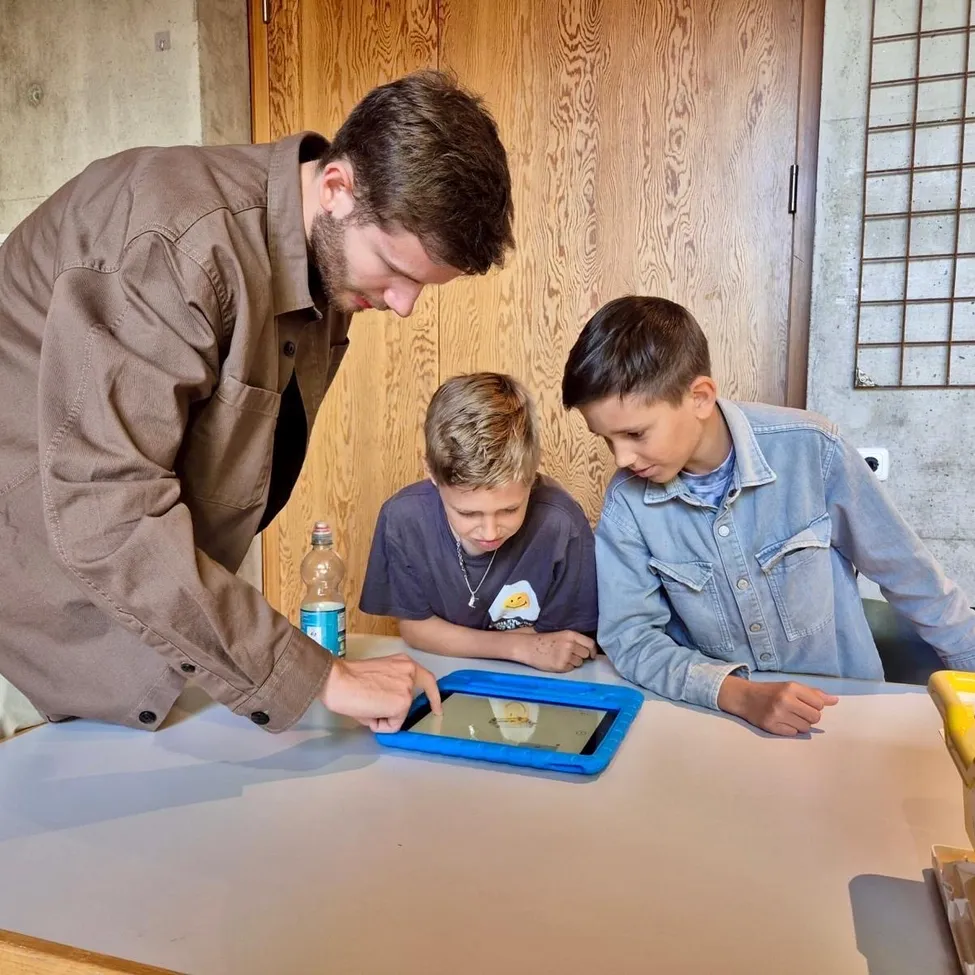 Ein Mann in einem braunen Hemd zeigt zwei Jungen in einem Klassenzimmer ein Tablet. Ein Junge trägt eine Jeansjacke und der andere ein blaues Hemd.