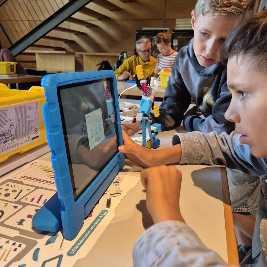 Eine Gruppe von Kindern sitzt um einen Tisch herum und arbeitet an einem Tablet mit einem Lego-Set auf dem Bildschirm. Sie konzentrieren sich auf die Aufgabe.