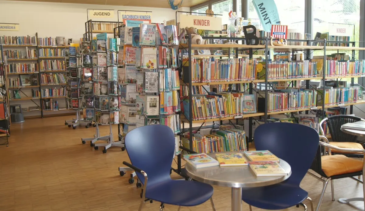 Bibliothek mit Regalreihen voller Bücher. Abschnitte mit den Bezeichnungen Jugend, Sachbuch Kinder und Kinder. Blaue Stühle um einen Tisch mit Büchern darauf.