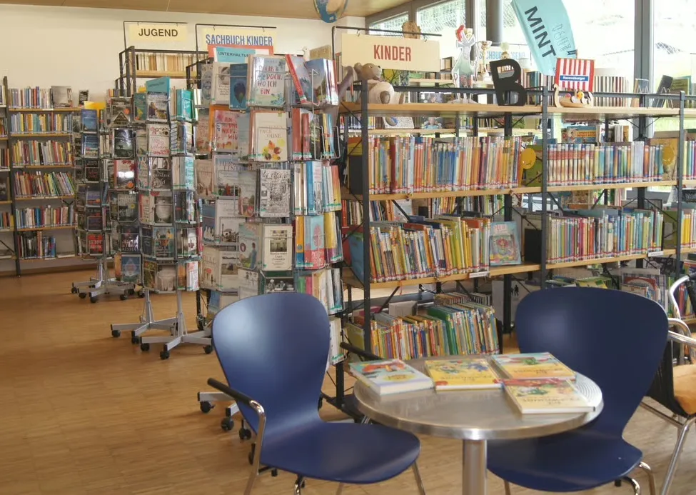 Bibliothek mit Regalreihen voller Bücher. Abschnitte mit den Bezeichnungen Jugend, Sachbuch Kinder und Kinder. Blaue Stühle um einen Tisch mit Büchern darauf.
