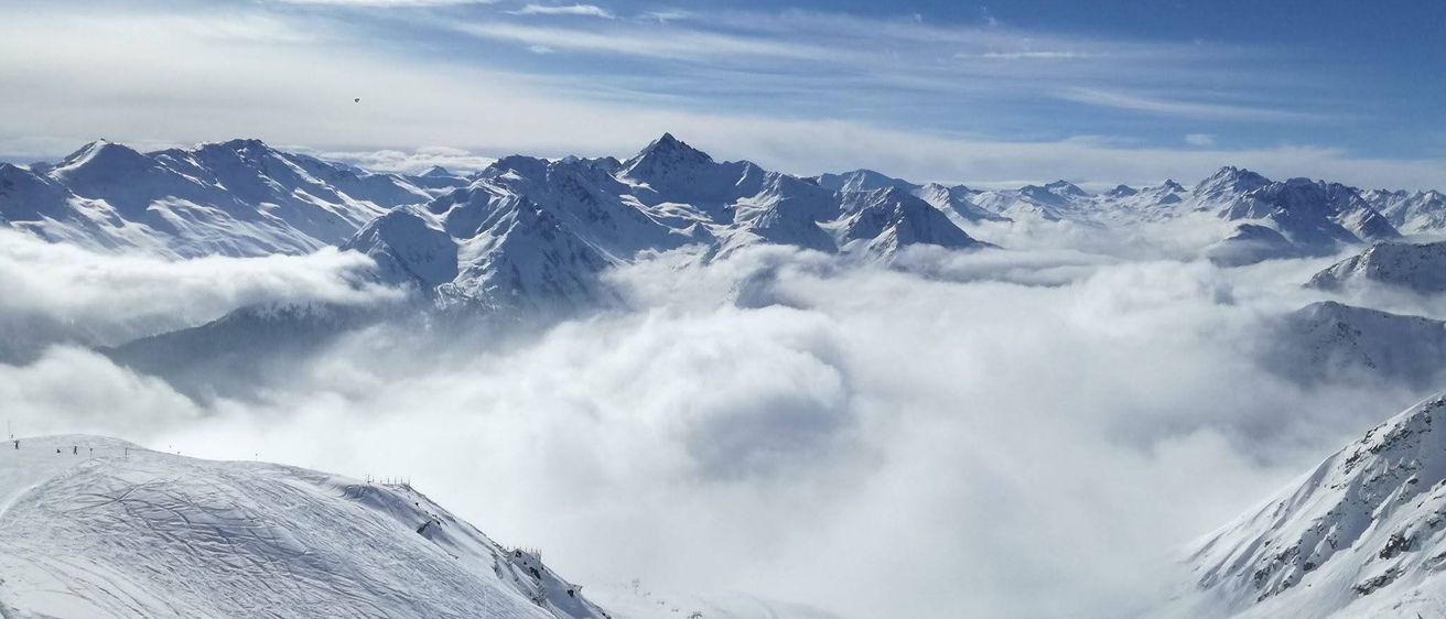 Luftaufnahme von schneebedeckten Bergen mit klarem blauem Himmel und verstreuten Wolken. Im Hintergrund sind Skilifte zu sehen.