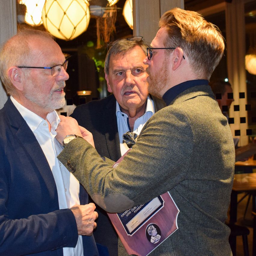 Drei Männer stehen in einem Raum. Einer trägt eine Brille und ein anderer hält eine Plakette. Sie scheinen sich bei einer Veranstaltung zu befinden.