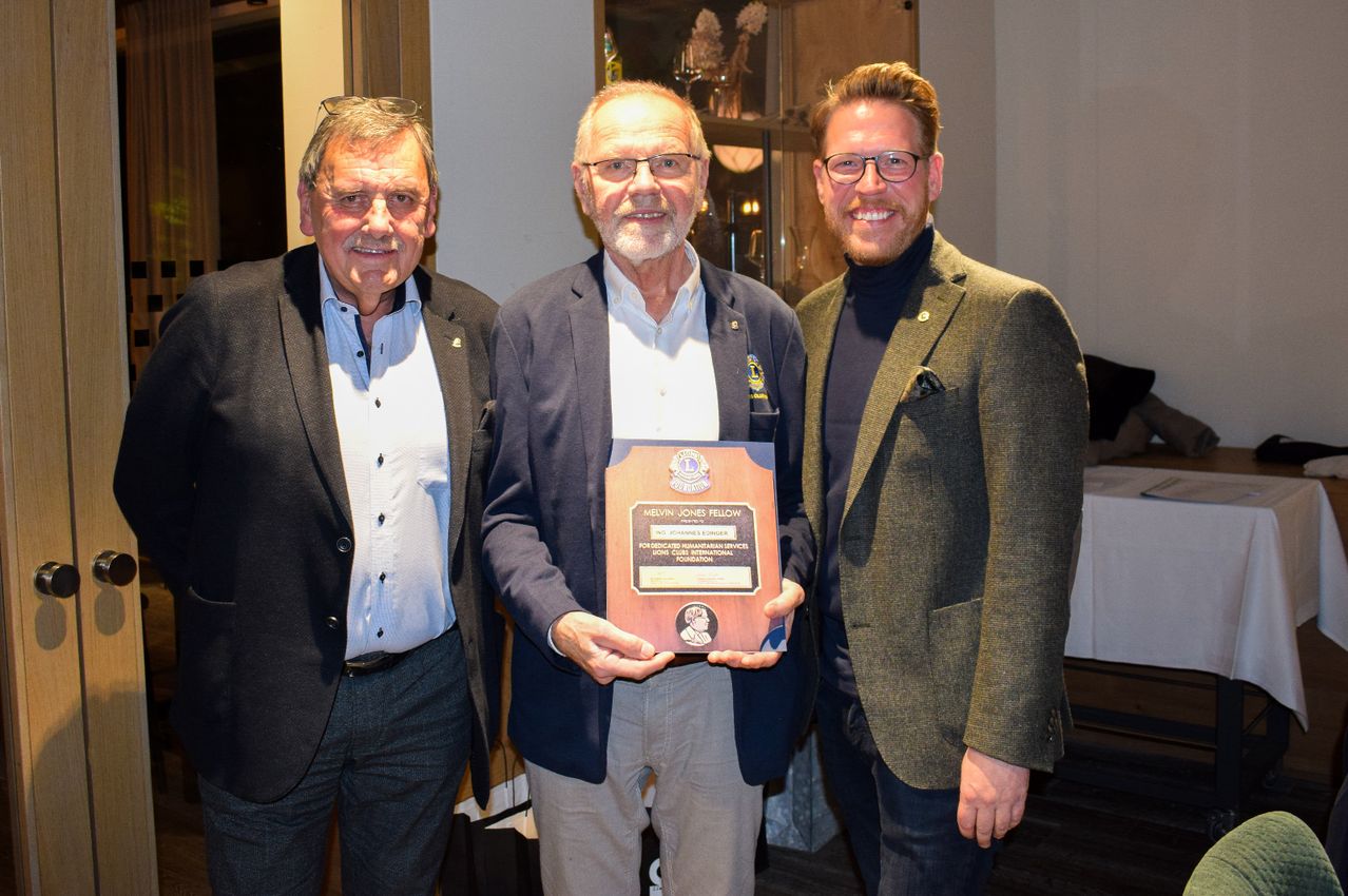 Drei Männer stehen zusammen und lächeln. Der Mann in der Mitte hält eine Plakette. Die Plakette hat ein Bild einer Person und einige Text darauf. Sie tragen formelle Kleidung. Hinter ihnen ist ein verschwommener Hintergrund.