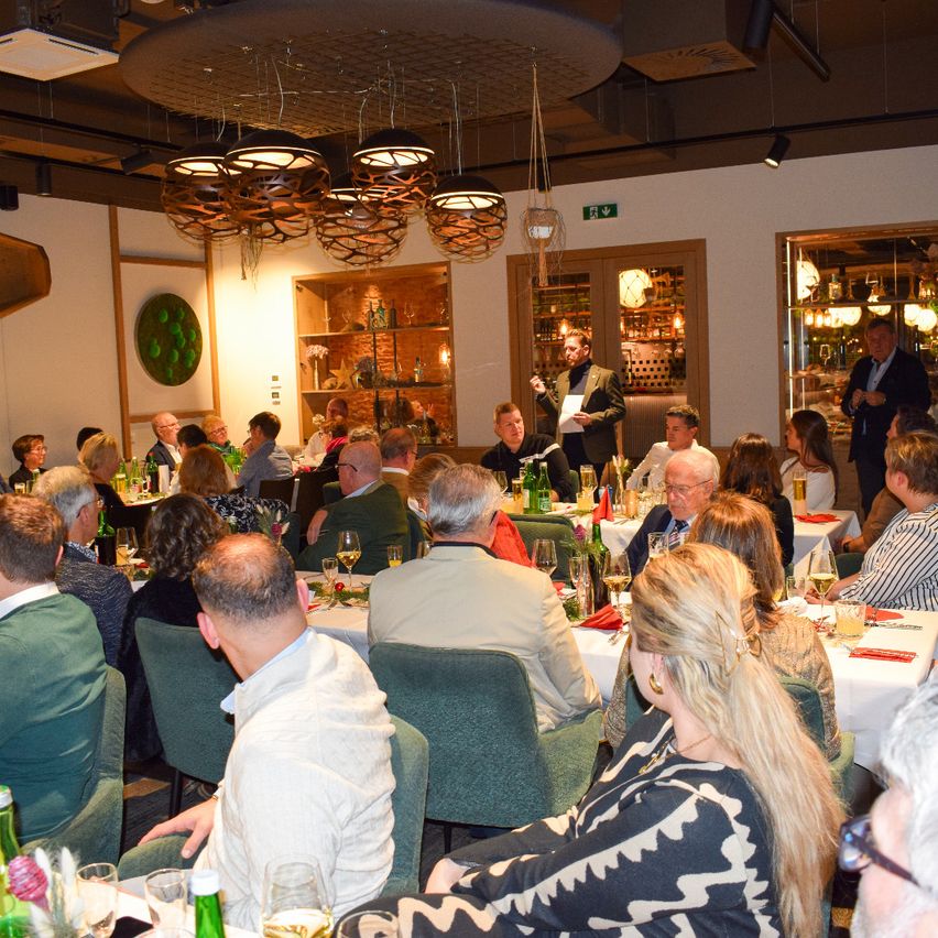 Eine Gruppe von Menschen sitzt an Tischen in einem Essbereich, einige hören einem Mann zu, der spricht. Die Tische sind mit Weingläsern, Flaschen und Servietten gedeckt. Der Raum hat ein modernes Design mit Lampen, die von der Decke hängen, und Regalen mit verschiedenen Gegenständen.