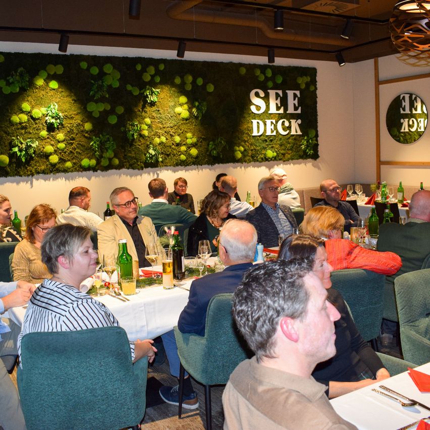 Eine Gruppe von Menschen sitzt um Tische in einem Restaurant mit einer Mooswand dahinter. Auf der Wand steht das Wort SEE DECK. Flaschen, Gläser und Teller stehen auf den Tischen.