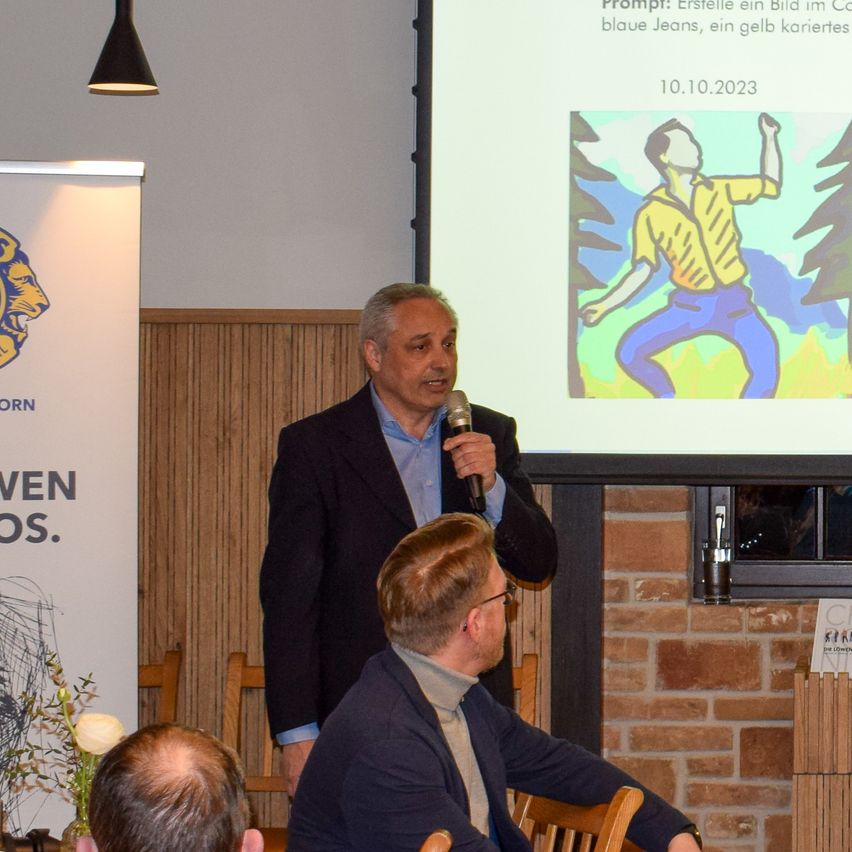 A man speaks into a microphone while another man sits in front of him. A poster with a lion logo is nearby. A projector screen displays a drawing.