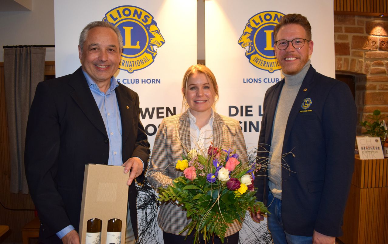 Three people stand in front of a backdrop with Lions Club Horn and Lions Club International logos. The man on the left holds a box with two wine bottles. The woman in the middle holds a bouquet of flowers and the man on the right smiles.