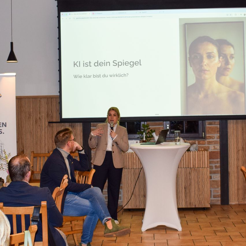 A woman stands at a podium in front of a projector screen displaying a picture of a woman with a serious expression and the words KI ist dein Spiegel. A man and woman sit at a table in front of her.