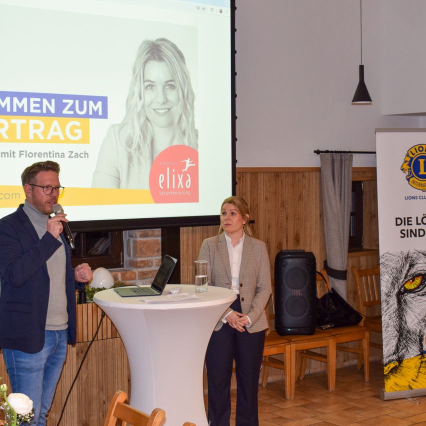 A man with a microphone stands beside a woman in a suit in a room. A projector screen displays a woman with the text 'ELIXA'. A speaker and a laptop sit on a table. A wooden chair and a wooden desk are on the right.