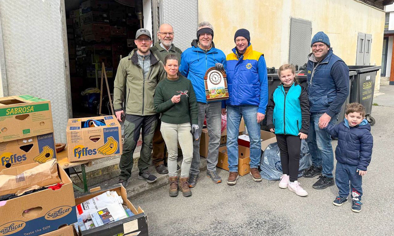 A group of people stand in front of a building. A woman holds a framed picture while a man in front holds a clock. Boxes and bags are around them.