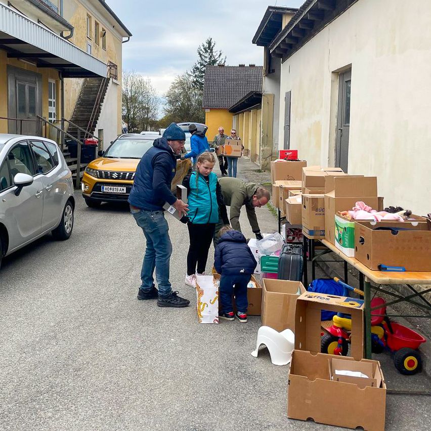 People gather in front of a building, sorting through boxes of donated items. A child inspects a box while a man looks over a table with toys.