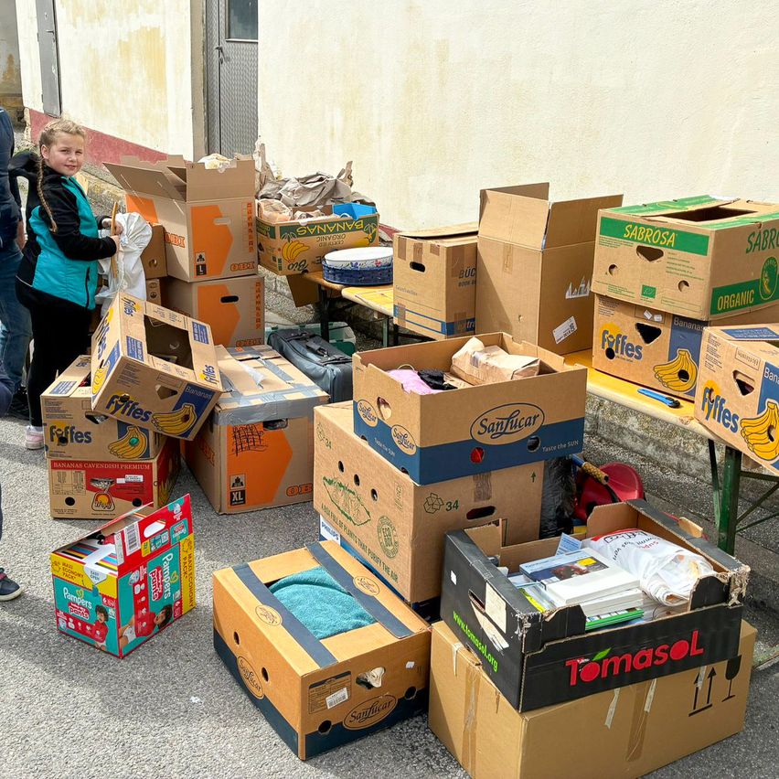 A child stands next to a pile of boxes, some containing bananas, others with items like diapers. The scene is outside, with a table and a building in the background.