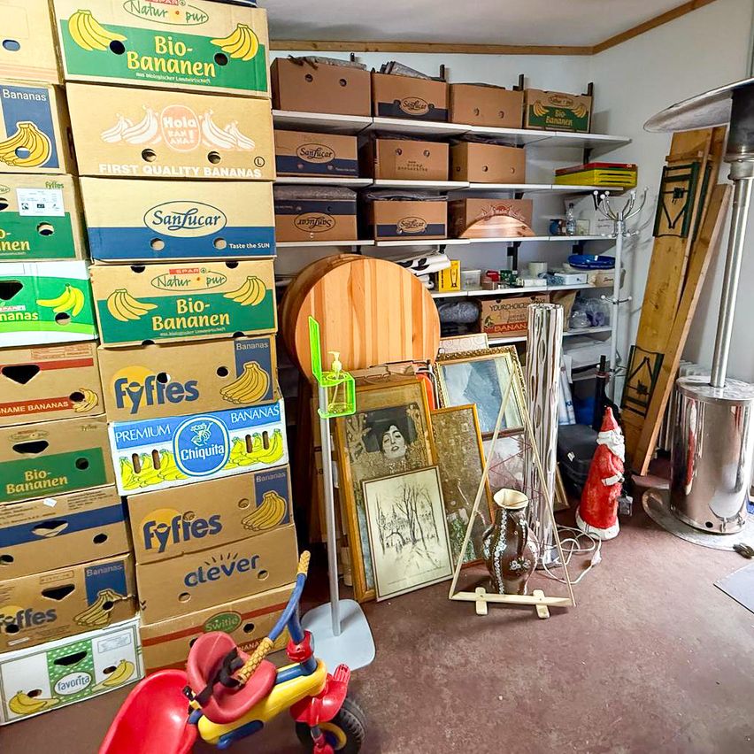 A room full of various boxes stacked, some of which are labeled with banana images. A few boxes are stacked on top of each other. A few picture frames are placed on the shelf. A red tricycle is placed in the room.