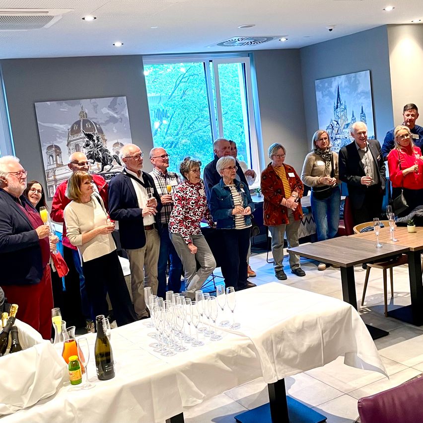 Eine Gruppe von Menschen versammelt sich in einem Raum, einige halten Weingläser, andere stehen in der Nähe von Tischen mit Flaschen und Gläsern. Kunstwerke schmücken die Wände, und natürliches Licht fällt durch Fenster ein.