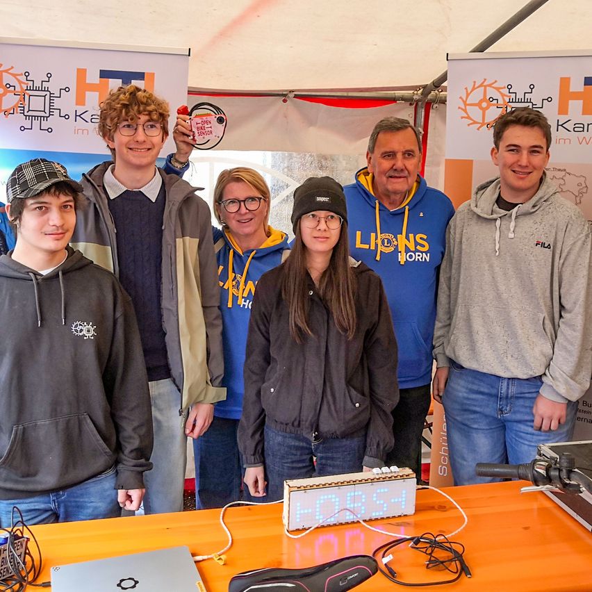 Eine Gruppe junger Personen in Hoodies steht hinter einem Tisch mit einem elektronischen Gerät, das '5085' anzeigt. Ein Banner mit einem Logo befindet sich hinter ihnen.