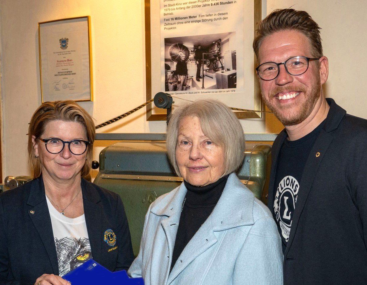 Drei Personen stehen eng beieinander in einem Raum. Eine Frau auf der linken Seite hält eine blaue Mappe, während die Frau in der Mitte einen Blazer trägt. Ein Mann auf der rechten Seite lächelt. Hinter ihnen ist eine Wand mit einem gerahmten Bild.