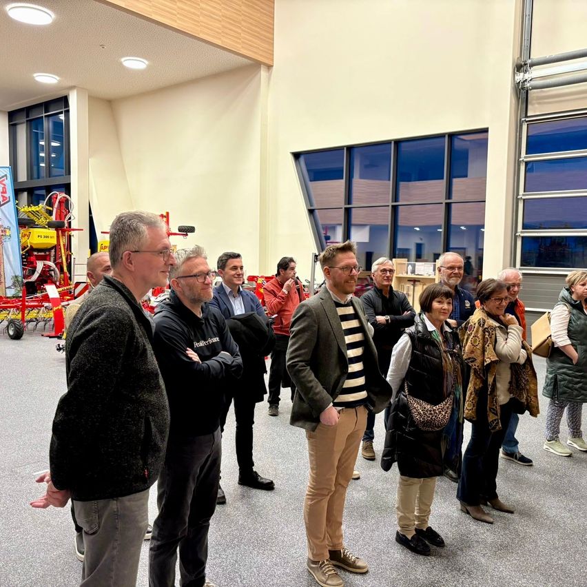 Eine Gruppe von Menschen steht in einem Raum, einige tragen Brillen und Turnschuhe, und zwei Männer vor ihnen tragen Mäntel und Hosen. Es gibt Glasfenster mit Blick nach draußen. Ein Maschinenfahrzeug ist auf der linken Seite geparkt, und ein Banner ist in der Nähe aufgestellt.