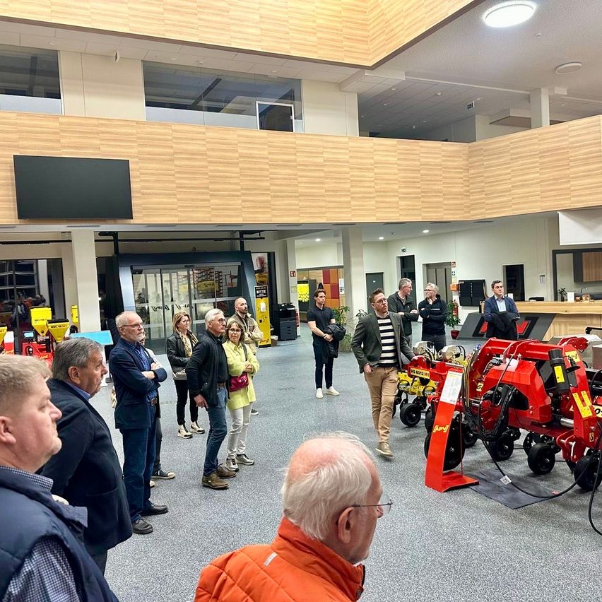 Eine Gruppe von Personen befindet sich in einem modernen Gebäude und untersucht rote Maschinen. Ein Mann in einer orangefarbenen Weste geht auf die Maschinen zu.