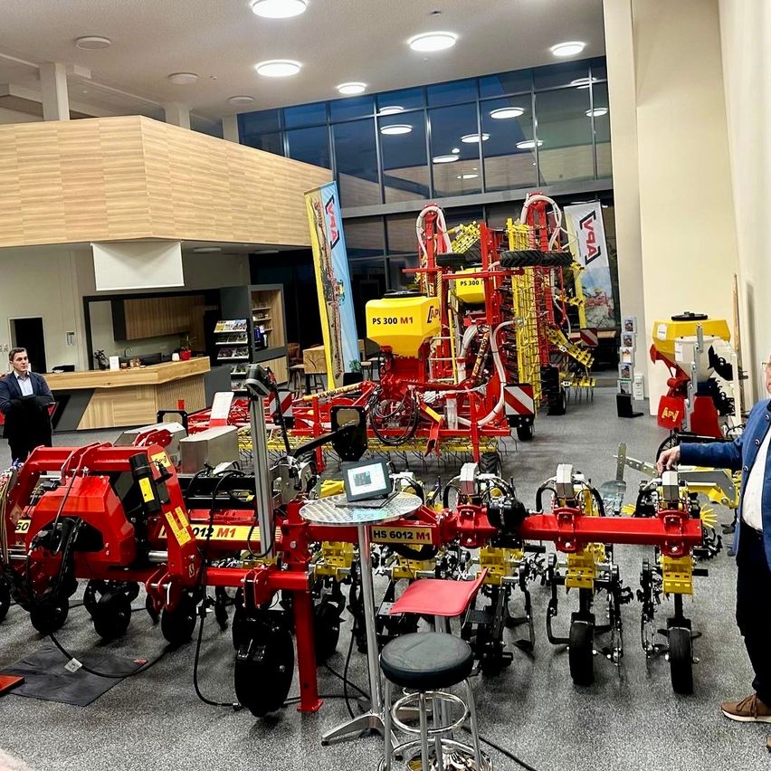 Eine Ausstellung im Innenbereich zeigt mehrere rote und gelbe landwirtschaftliche Maschinen, wobei ein Mann in einem blauen Anzug eine untersucht. Im Hintergrund befinden sich Banner und Lichter.