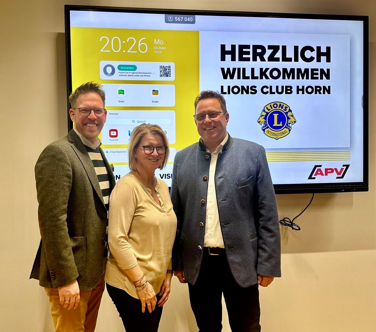 Drei Personen stehen vor einem Bildschirm mit einer Willkommensbotschaft für den Lions Club Horn. Der Bildschirm zeigt die Zeit und einen QR-Code an. Die beiden Männer tragen Mäntel und die Frau trägt eine Brille und eine Halskette.