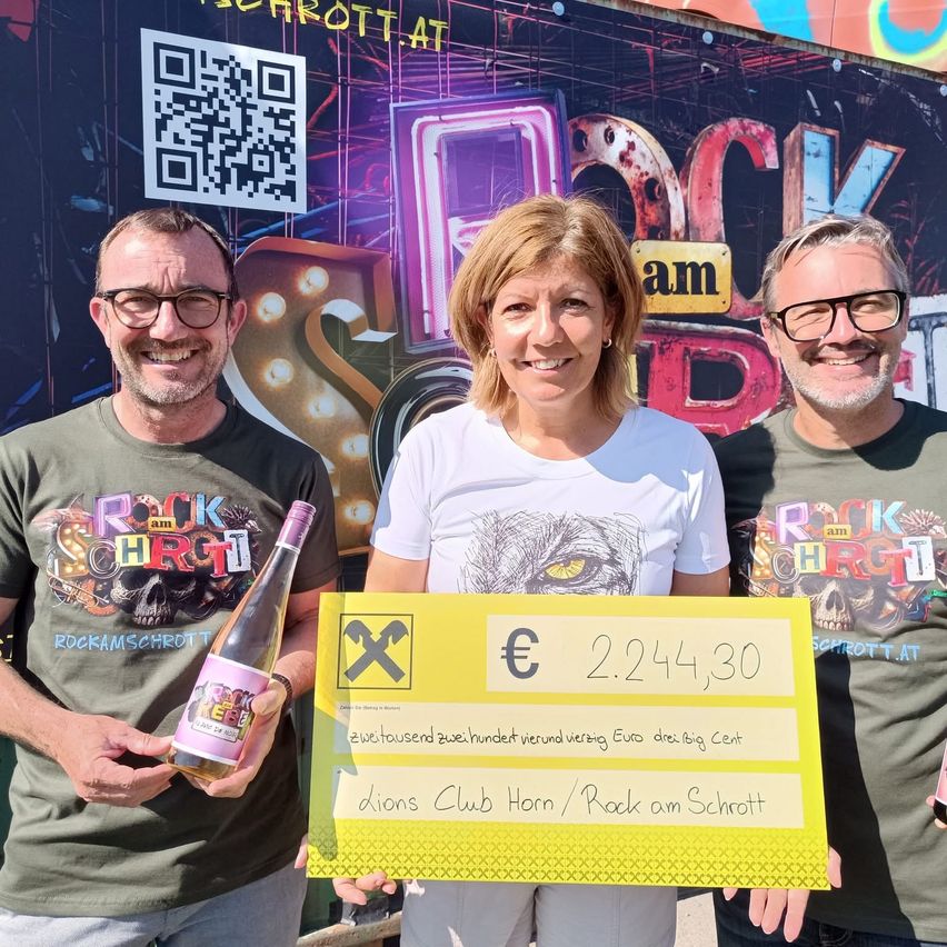 Drei Personen stehen vor einer bunten Wand mit einem QR-Code und lächeln. Die Frau in der Mitte hält einen gelben Scheck über 2.244,30 Euro. Der Mann auf der linken Seite hält eine Flasche Wein.