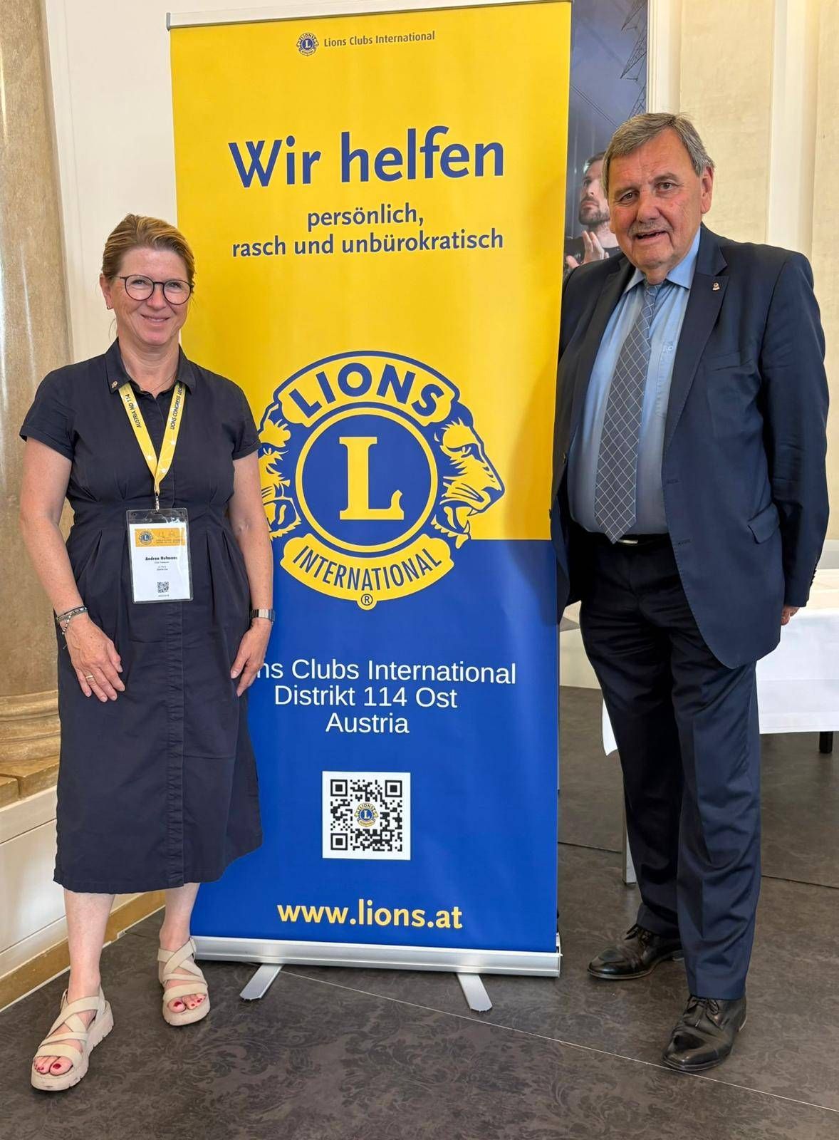 A woman and a man stand next to a blue banner with the text Lions International. The woman wears glasses and a badge. The man wears a suit and tie.