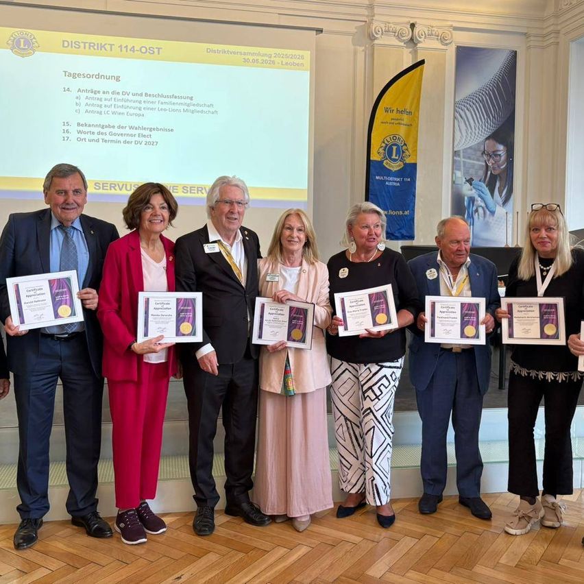 A group of people stand together holding awards in front of a projector screen. They wear formal clothing. The room has wooden flooring and banners.
