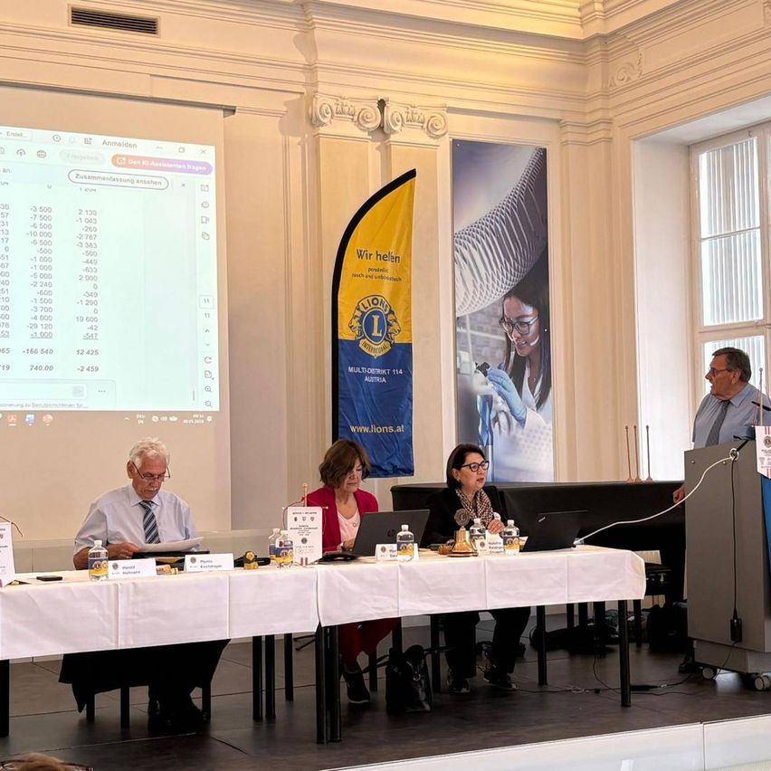 A group of people are sitting at a table during a presentation. A man at a podium speaks to them. A projector screen displays a slide. A banner is visible in the background.