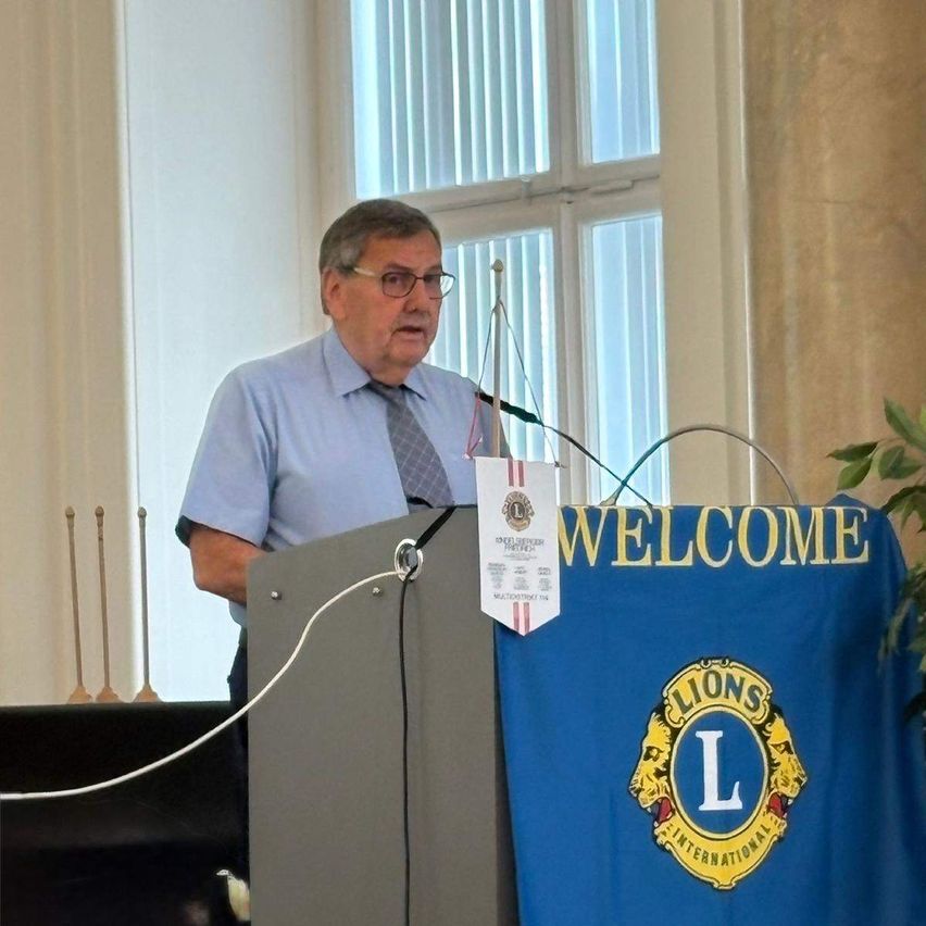 A man in a blue shirt and tie stands at a podium with a microphone, with a blue backdrop reading 'Welcome' and a Lions International logo.