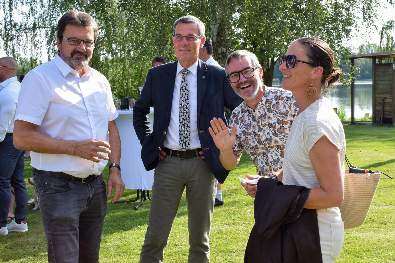 A group of people, including men and women, are standing in an outdoor area. They are all smiling and seem to be having a conversation. Behind them are trees and a table with glasses.