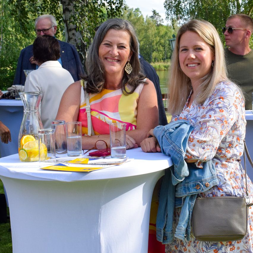 Two women smiling at a table with drinks and lemons, surrounded by others in a park.