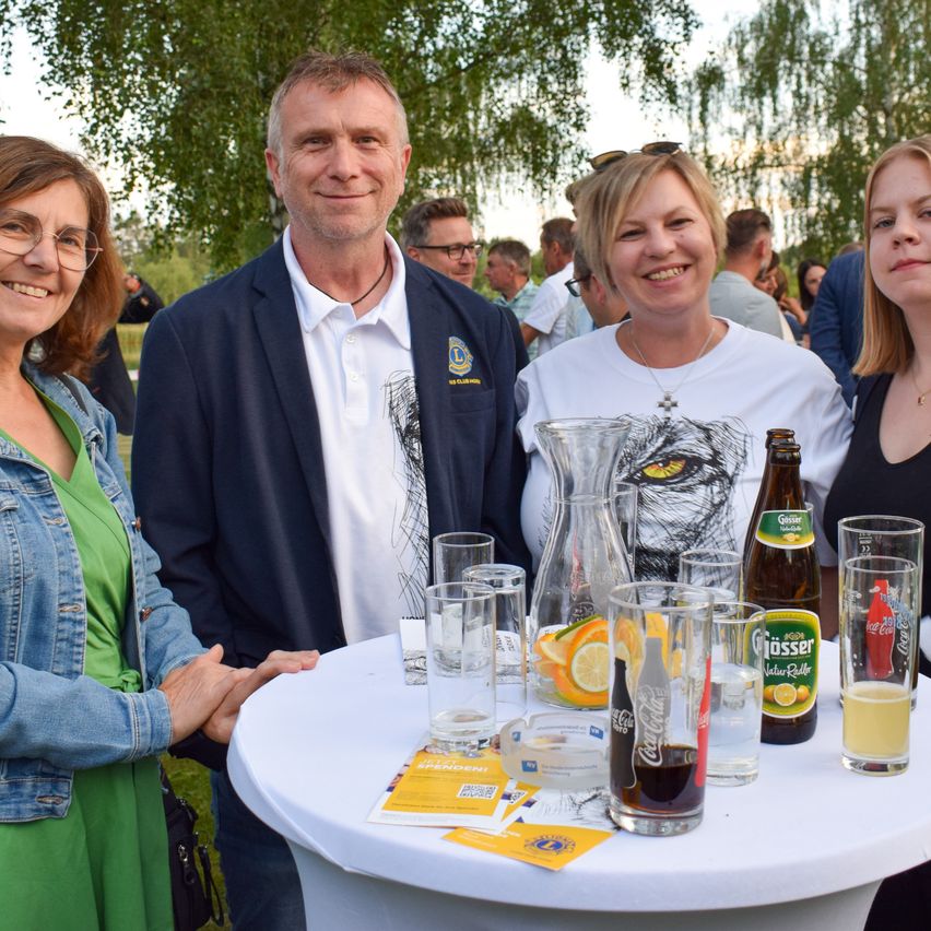 Four individuals stand around a table with drinks and food. One man wears a white shirt with a graphic, while others smile. The table has glasses, bottles, and a pitcher.