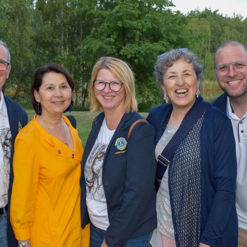 Five people stand in a group, smiling. They are dressed in various colors, with trees and a body of water in the background.