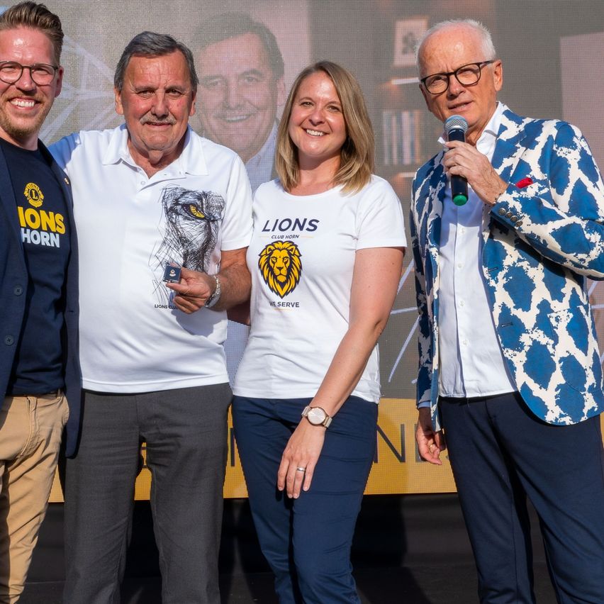 Vier Personen stehen auf einer Bühne vor einer großen Leinwand. Ein Mann hält ein Mikrofon und spricht. Eine Frau hält eine kleine Auszeichnung. Die anderen tragen Lions Club-Shirts.