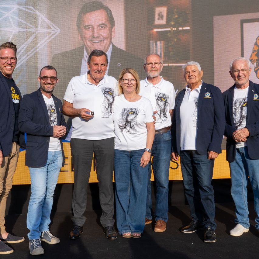 Eine Gruppe von Menschen steht vor einer Kulisse, mit einem lächelnden Mann auf dem Bildschirm. Sie tragen passende Shirts und posieren für ein Foto.