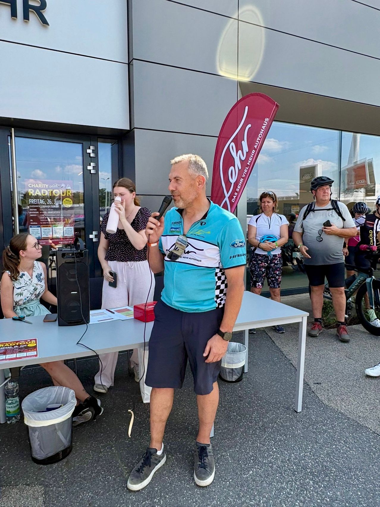 Ein Mann spricht in ein Mikrofon bei einer Outdoor-Veranstaltung. Hinter ihm trinkt eine Frau aus einer Flasche. Ein Banner und Fahrräder sind im Hintergrund.