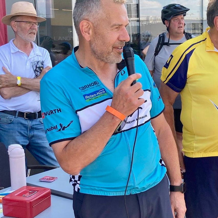 Ein Mann in einem Radtrikot spricht in ein Mikrofon, ein weiterer Mann mit Helm steht neben ihm, ein weiterer Mann in einem gelben Shirt beobachtet.