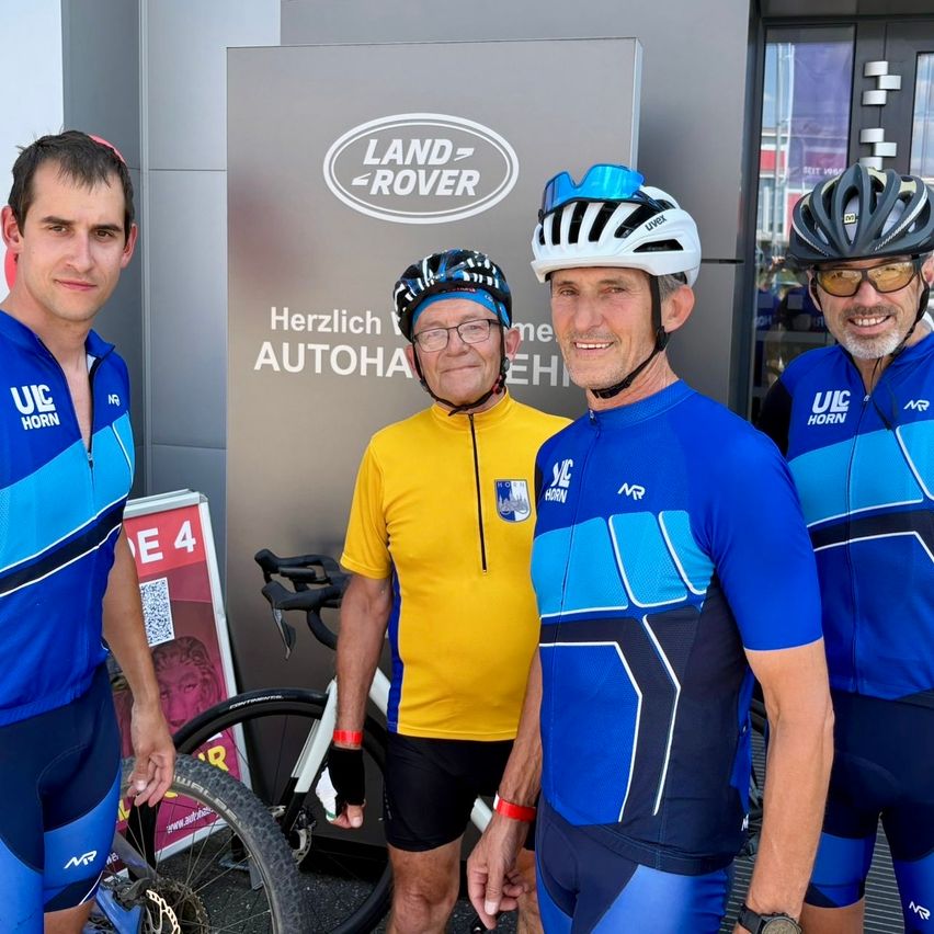 Vier Männer in Radfahrerkleidung stehen vor einem Gebäude mit dem Schild 'Land Rover'. Sie alle lächeln und scheinen für ein Foto zu posieren.