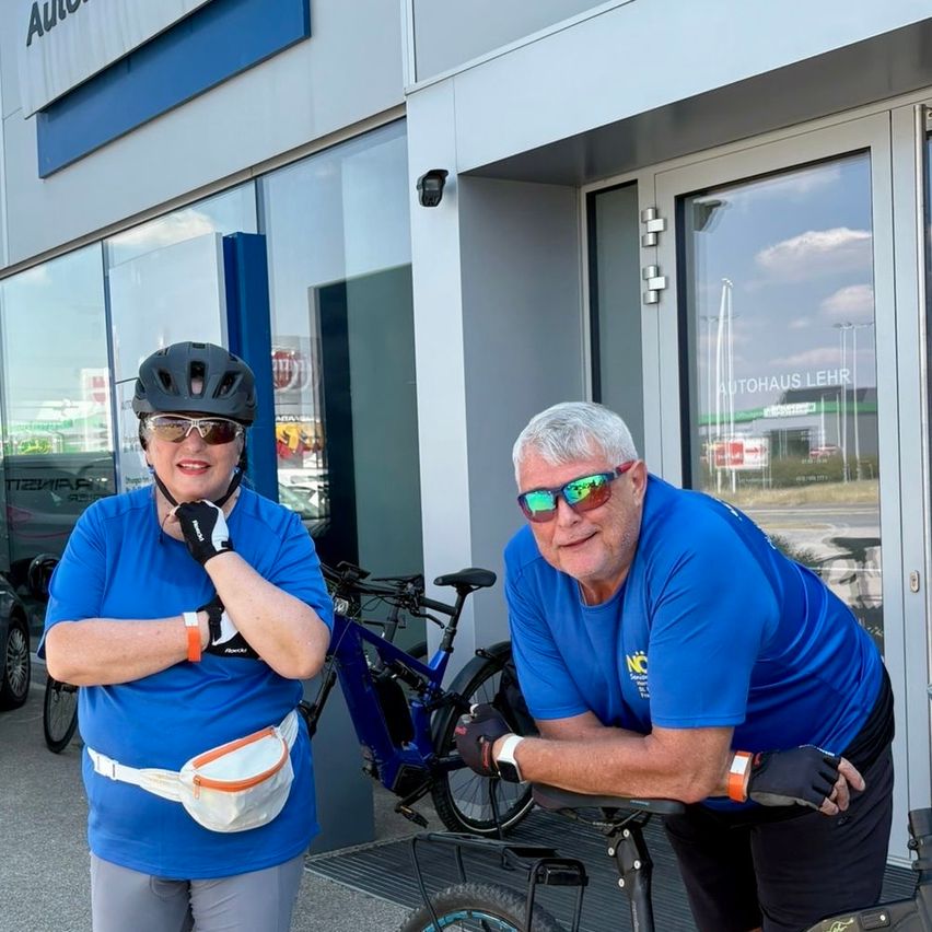 Zwei Menschen in blauen Hemden und Helmen posieren mit Fahrrädern vor einem Gebäude mit AUTOHAUS LEHR an der Tür.