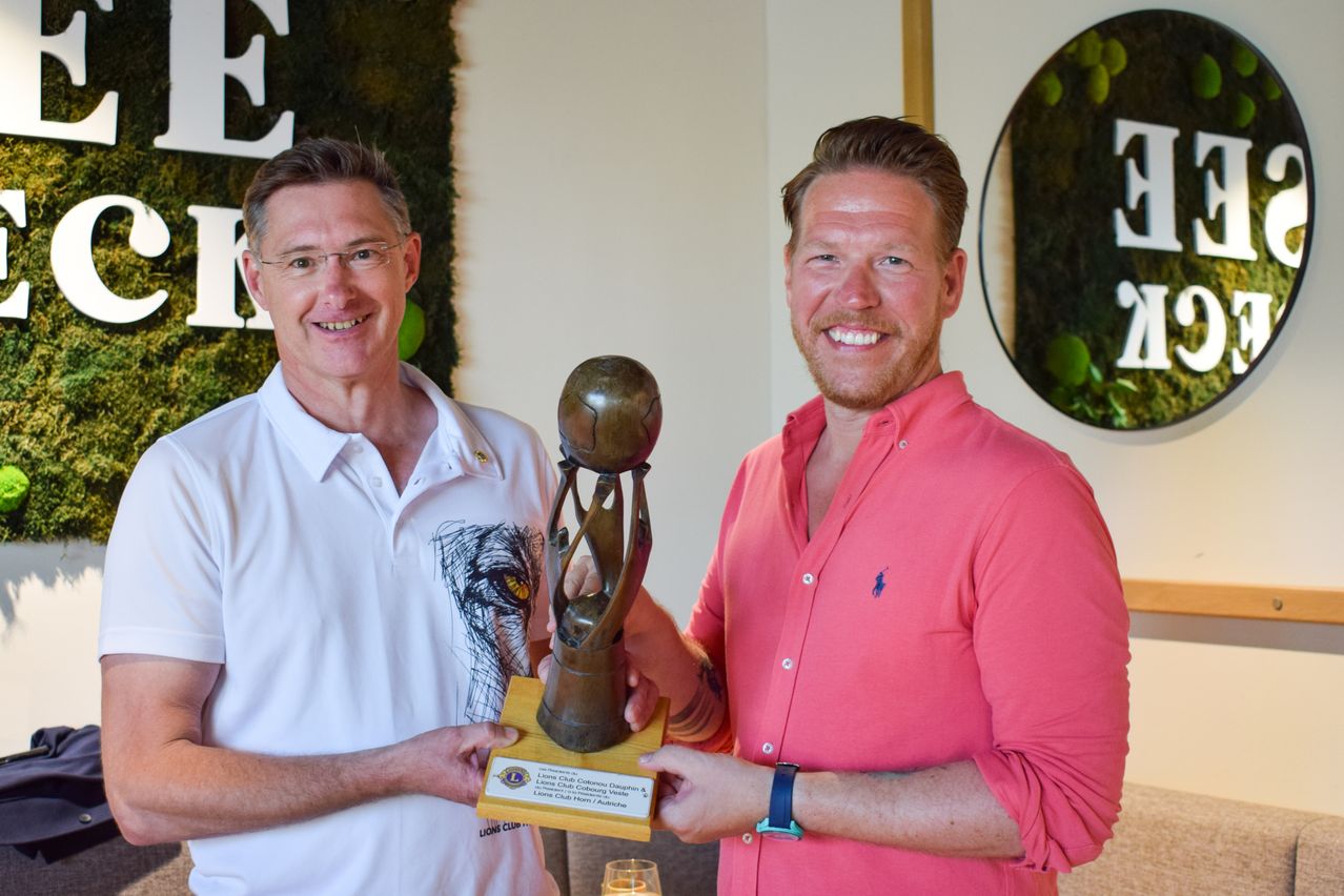 Zwei Männer lächeln und posieren mit einer Trophäe in einem Raum. Ein Mann trägt ein weißes Poloshirt, der andere ein rosa Hemd. Die Trophäe ist bronzefarben und hat eine Weltkugel obenauf. Die Wand hinter ihnen hat ein grünes Wanddesign und einen Spiegel.